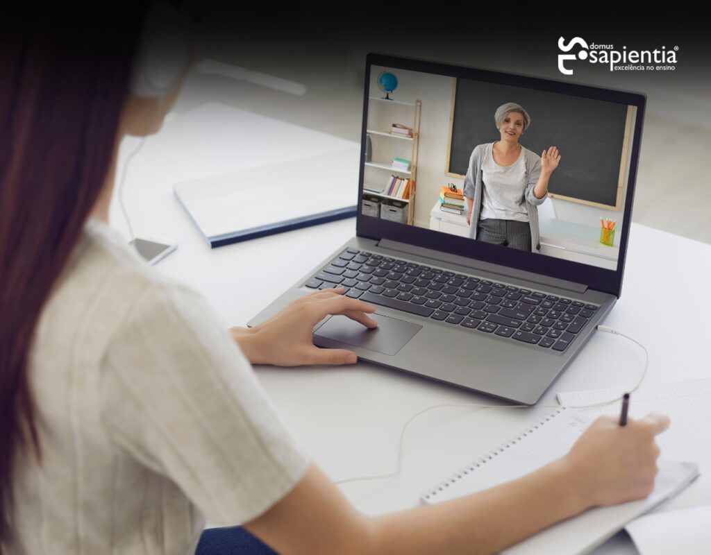 Uma pessoa está sentada em uma mesa participando de uma palestra online em um laptop. Um professor é visível na tela do laptop gesticulando em frente a um quadro branco. Cadernos e caneta estão na mesa.