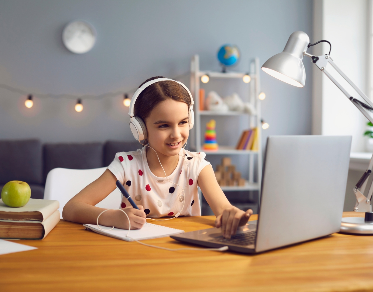 Uma garota usando fones de ouvido está sentada em uma mesa, olhando para um laptop. Ela está escrevendo em um caderno. A mesa tem livros, uma luminária e uma maçã verde. O fundo mostra prateleiras com decorações.