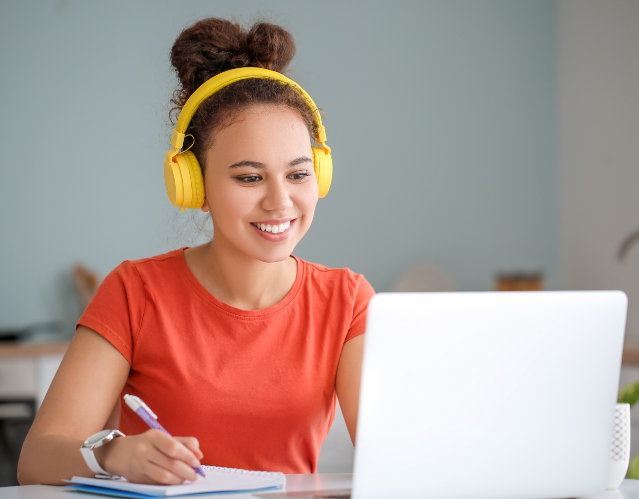 Uma pessoa com cabelos cacheados presos em um coque, usando fones de ouvido amarelos e uma camisa vermelha, sorri enquanto trabalha em um laptop e escreve em um caderno.