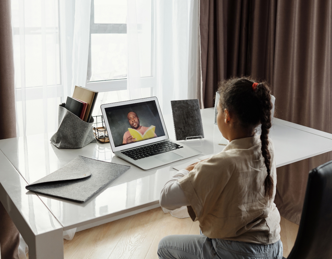 Uma criança está sentada em uma mesa, participando de uma sessão de aprendizado virtual em um laptop com um professor lendo um livro na tela. A mesa está organizada com pastas e um porta-canetas.