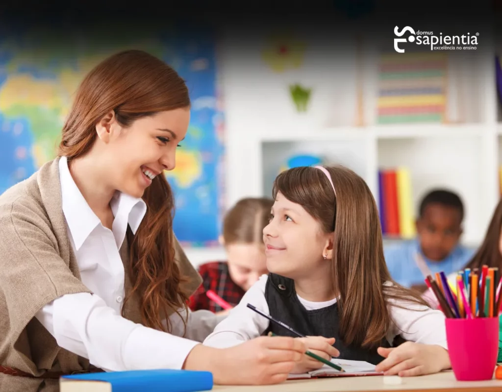Um professor e um aluno sorriem um para o outro em uma sala de aula. O professor está sentado ao lado do aluno, que segura um lápis. Há livros e materiais coloridos na mesa, e outros alunos ao fundo.