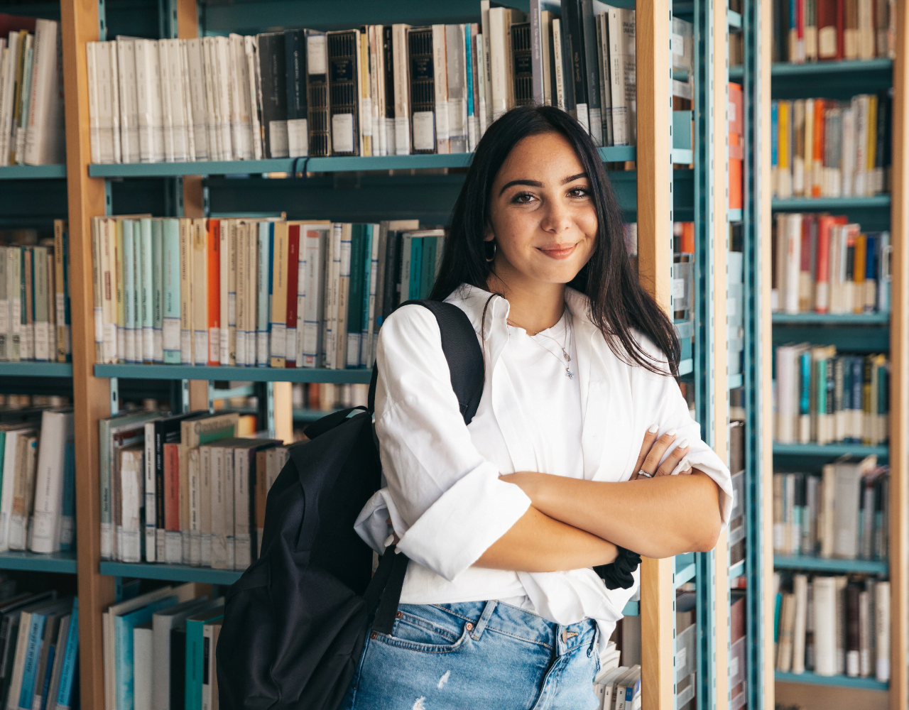 Uma jovem parada em uma biblioteca com os braços cruzados.