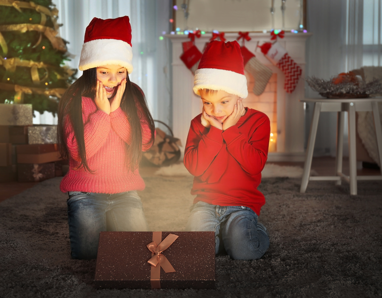 Duas crianças com chapéus de Papai Noel sentadas no chão em frente a uma árvore de Natal, comemorando o Natal 2023.