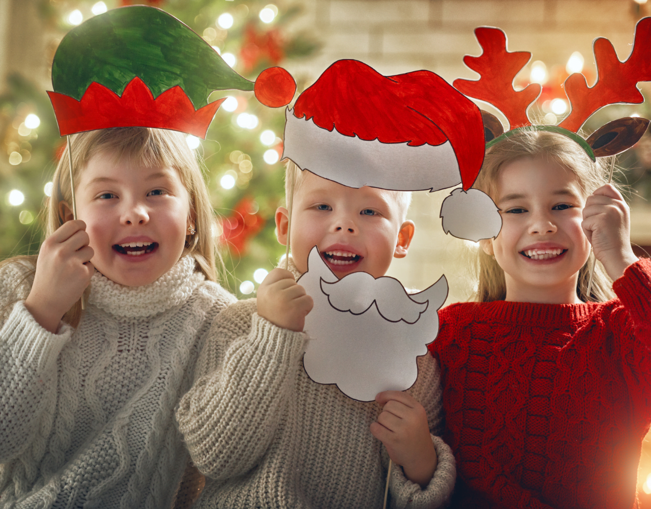 Três crianças segurando recortes de papel de Papai Noel em frente a uma árvore de Natal.