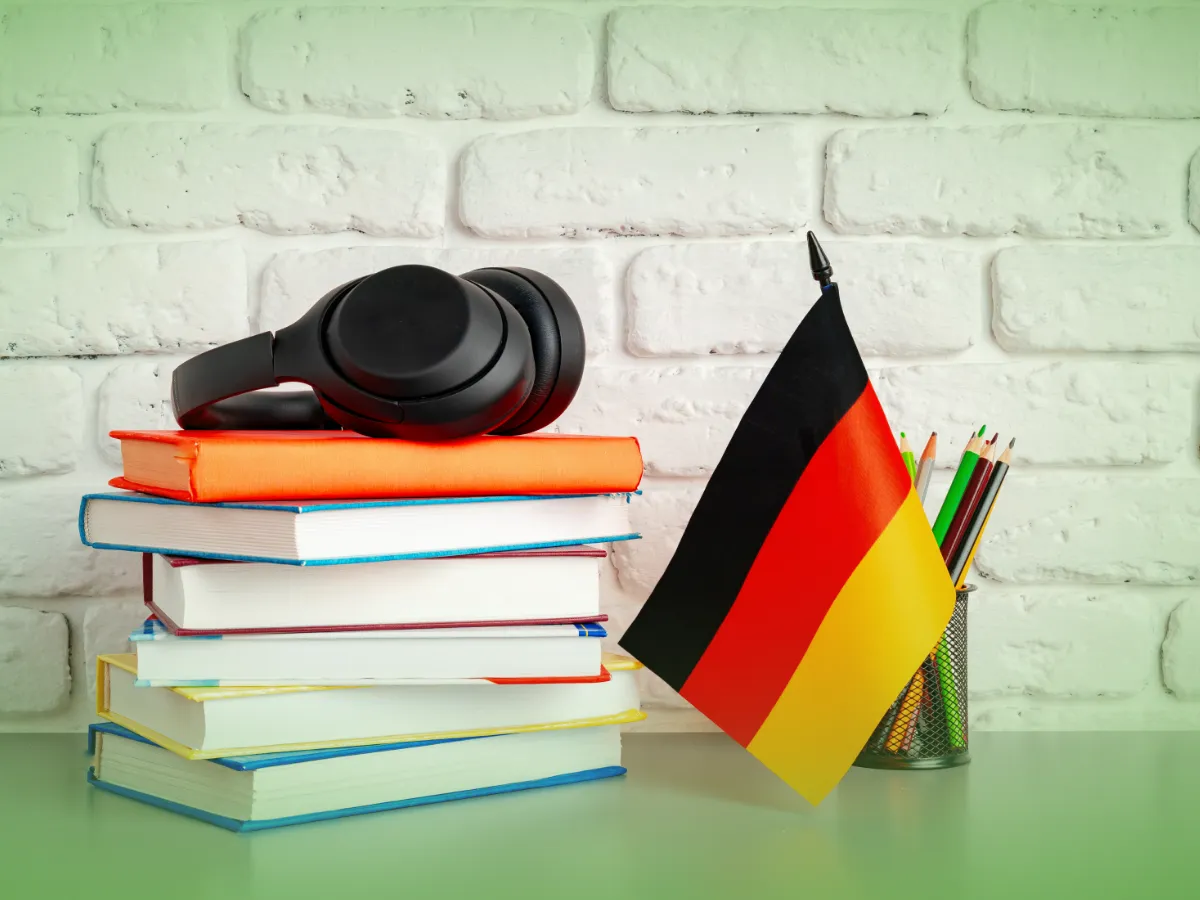 Uma pilha de livros coloridos de Explicações do Alemão 10.º Ano com fones de ouvido pretos no topo, ao lado de uma bandeira alemã e um porta-lápis com lápis de cores variados sobre um fundo de tijolo branco.