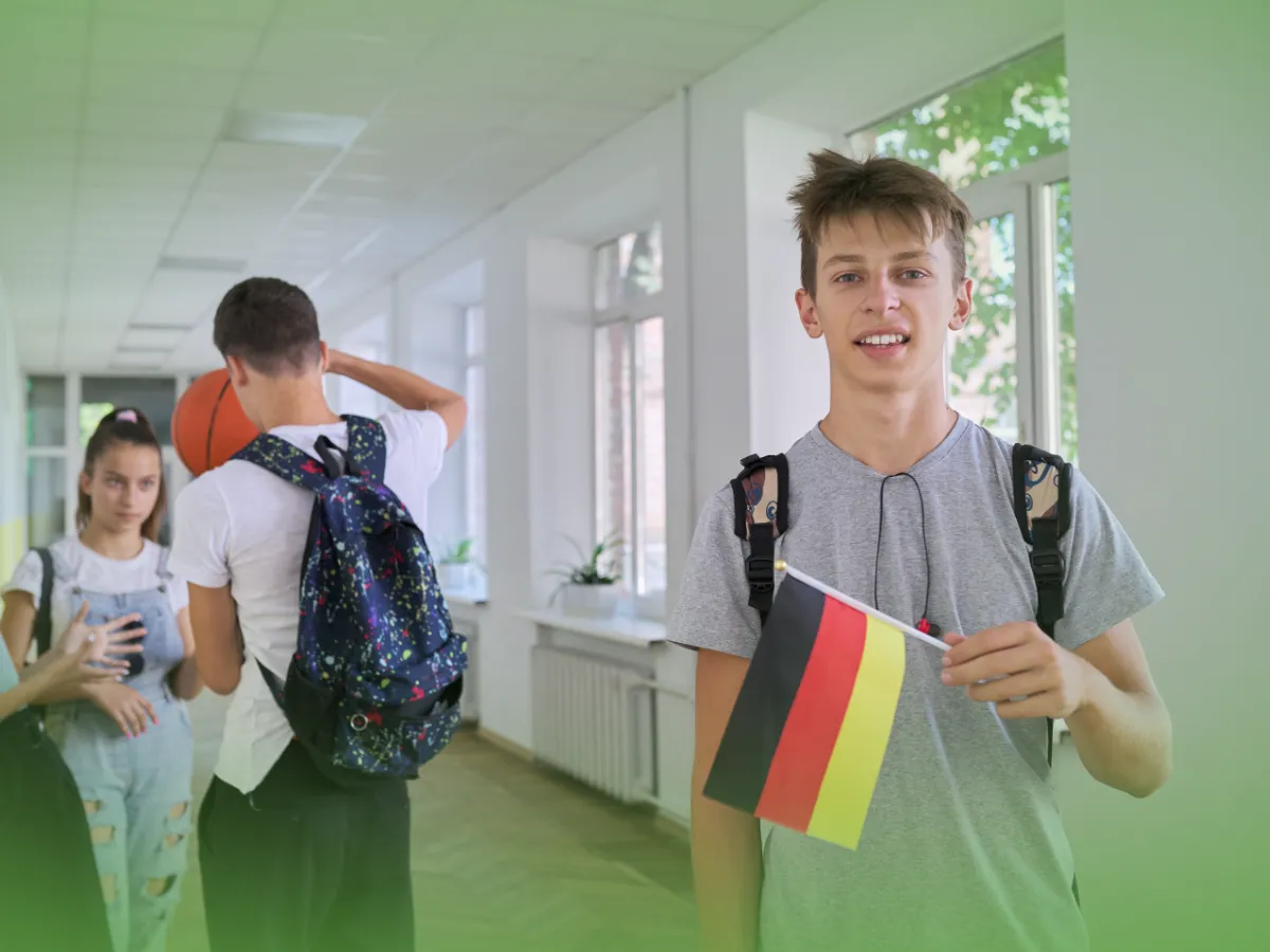 Menino adolescente com mochila segurando Explicações de Alemão 8.º Ano - Pacote de Horas no corredor de uma escola, com outros alunos ao fundo.