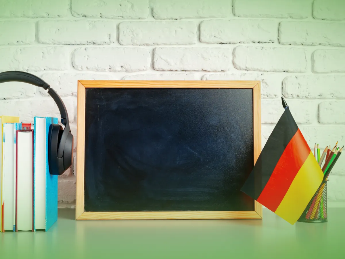 Um quadro-negro, livros, fones de ouvido e um porta-lápis sobre uma mesa encostada em uma parede de tijolos brancos com Explicações de Alemão 9.º Ano. Pacote de Horas.