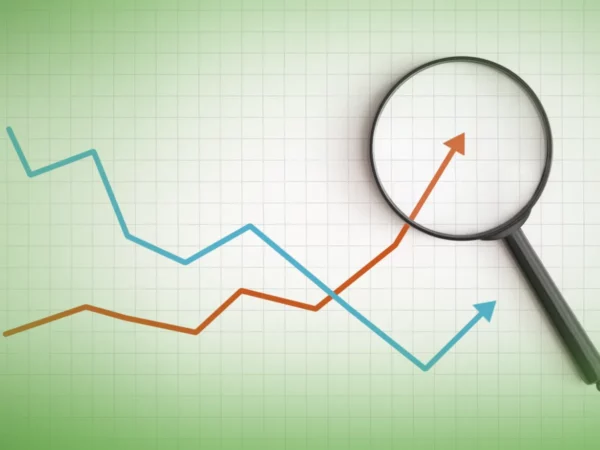 Uma lupa paira sobre uma grelha com dois gráficos de linhas que se cruzam, um azul e outro vermelho, mostrando tendências flutuantes de dados para Explicações de Economia A 11.º Ano. Pacote de Horas.