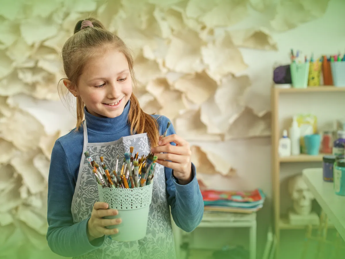 Uma jovem sorrindo ao selecionar Explicações de Educação Visual 6.º Ano. Pacote de Horas em um porta-pincéis diversos, em uma sala colorida e artística.
