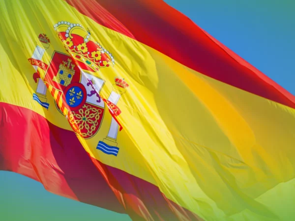 Uma bandeira vibrante das Explicações de Espanhol 10.º Ano, com um brasão detalhado, ondulando contra um céu azul claro.