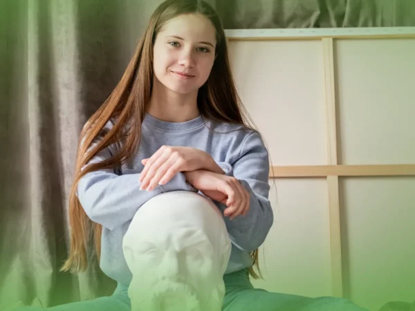 Jovem de cabelos longos sorrindo para a câmera, sentada atrás de uma escultura branca de uma cabeça colocada sobre uma mesa, com cortinado verde ao fundo de Explicações de Filosofia 10.º Ano. Pacote de Horas.