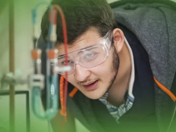 Uma pessoa usando óculos de segurança observa uma experiência científica com vários tubos e fios usando Explicações de Física 12.º Ano. Pacote de Horas.