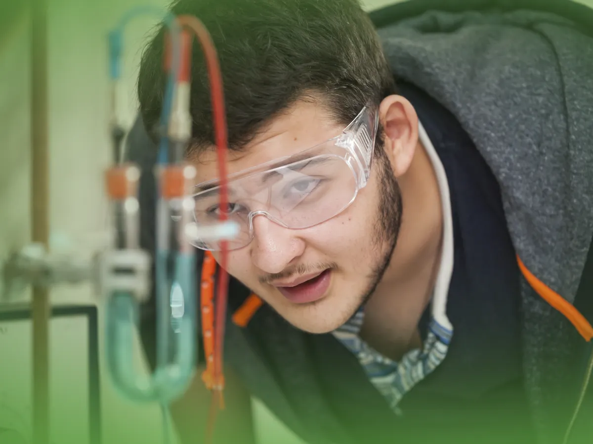 Uma pessoa usando óculos de segurança observa uma experiência científica com vários tubos e fios usando Explicações de Física 12.º Ano. Pacote de Horas.