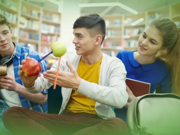 Três alunos sentados numa biblioteca, um deles explicando um conceito usando Explicações de Física e Química A 10.º Ano. Pacote de Horas e uma modelo enquanto as outras duas ouvem atentamente.