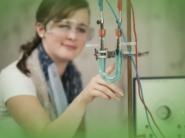 Uma pessoa usando óculos de segurança ajusta um aparato científico envolvendo tubos e fios de vidro em ambiente de laboratório usando as Explicações de Física e Química A 11.º Ano. Pacote de Horas.