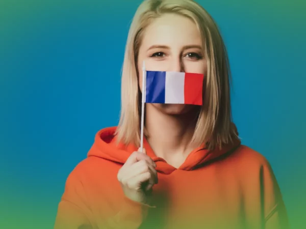 Uma pessoa com um moletom laranja segura uma pequena bandeira francesa na frente da boca, contra um fundo azul. Explicações de Francês 11.º Ano . Pacote de Horas.