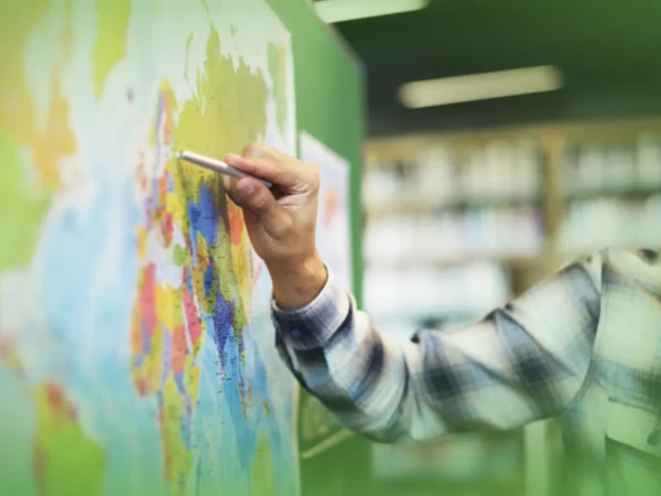 Uma pessoa de camisa xadrez apontando com um lápis para um mapa-múndi colorido, focando em detalhes geográficos específicos das "Explicações de Geografia 9.º Ano. Pacote de Horas".