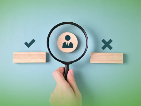 Mão segurando uma lupa sobre um ícone de uma pessoa entre uma marca de seleção e uma cruz em blocos de madeira contra um fundo verde. Explicações de Gestão de Recursos Humanos ONU . Pacote de Horas.