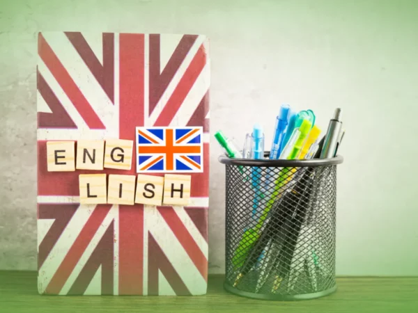 A Explicações de Inglês 11.º Ano . Pacote de Horas com capa da bandeira britânica e “Inglês” escrito em letras, ao lado de um porta-canetas cheio de canetas coloridas sobre uma escrivaninha.