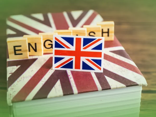 Um livro com capa Union Jack tem a palavra "ENGLISH" escrita em blocos de letras de madeira, com uma pequena bandeira da Union Jack na frente intitulada "Explicações de Inglês 12.º Ano. Pacote de Horas".