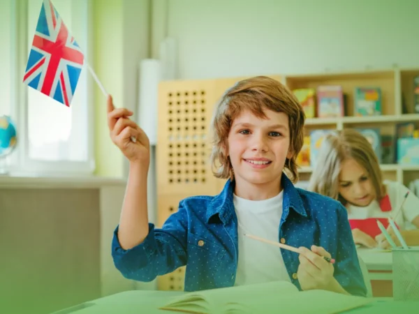 Um menino na sala de aula sorri enquanto segura um pequeno Explicações de Inglês 6.º Ano. Bandeira do Pacote de Horas, com uma garota focada em artesanato ao fundo.