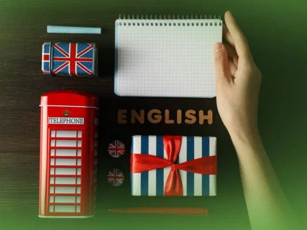 Uma mão abre um bloco de notas sobre uma mesa com itens de temática britânica: modelo de cabine telefônica, bandeiras, caixa embrulhada para presente e a palavra "Explicações de Inglês 7.º Ano . Pacote de Horas" em letras de madeira.