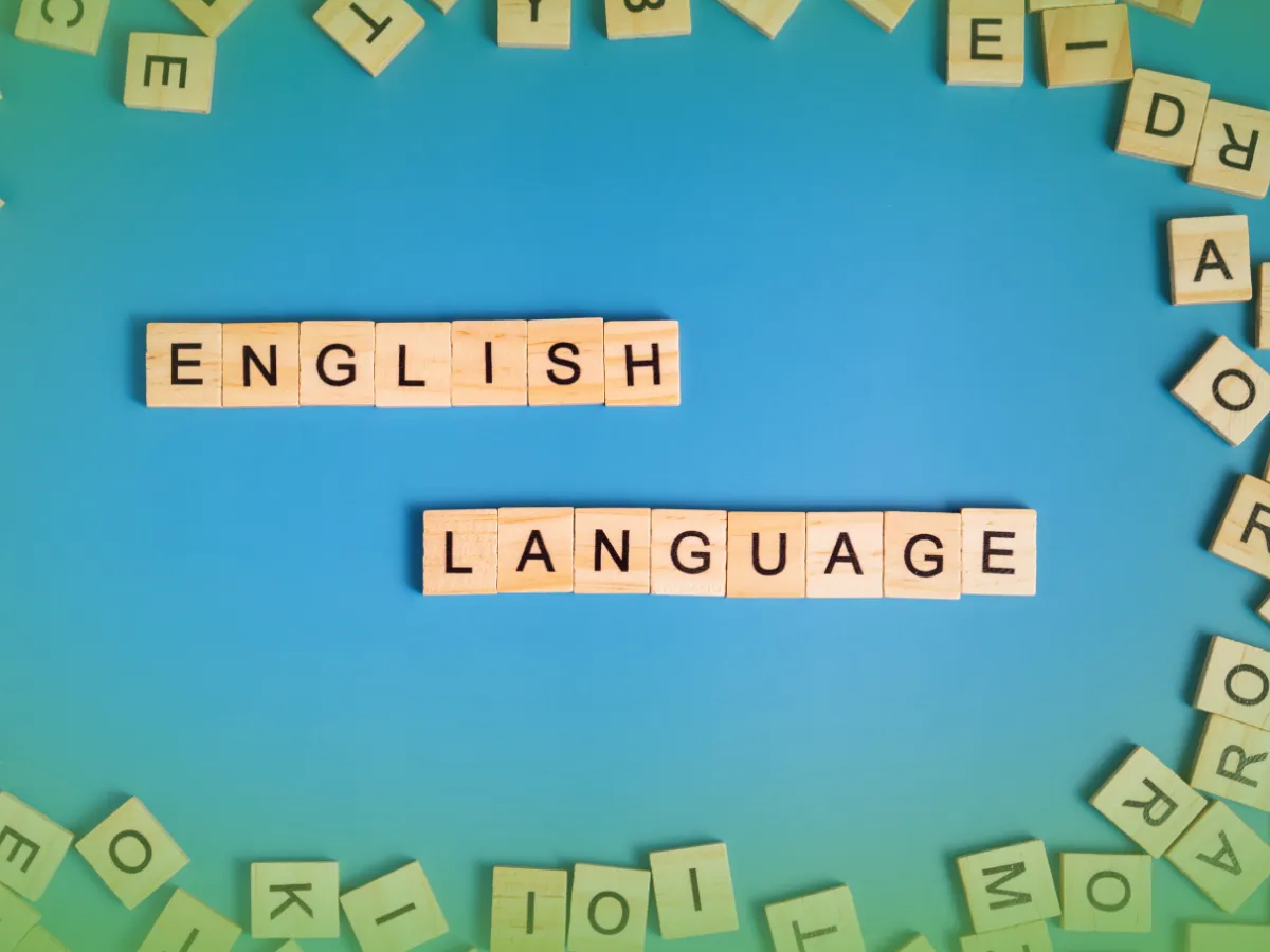 Blocos de madeira com a grafia "Explicações de Inglês 9.º Ano . Pacote de Horas" rodeados por ladrilhos de alfabeto espalhados sobre fundo azul.