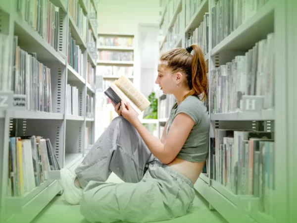Uma jovem em traje casual está sentada no chão entre as estantes da biblioteca, lendo Explicações de Língua Portuguesa 11.º Ano. Pacote de Horas atentamente.