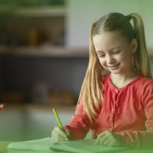 Uma jovem com rabo de cavalo, camisa vermelha, sorri enquanto desenha em papel verde numa mesa com lápis de cor próxima. Explicações Língua Portuguesa 2.º Ano . Pacote de Horas.