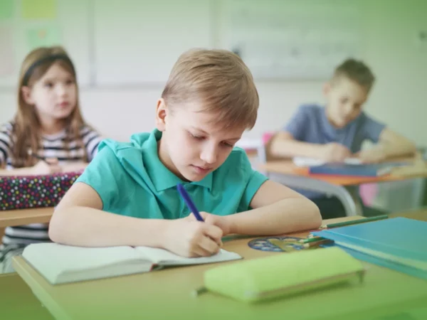 Frase com Nome do Produto: Um menino de camisa azul-petróleo escrevendo em um caderno na mesa da sala de aula, concentrado, com dois colegas ao fundo usando Explicações de Língua Portuguesa 3.º Ano. Pacote de Horas.