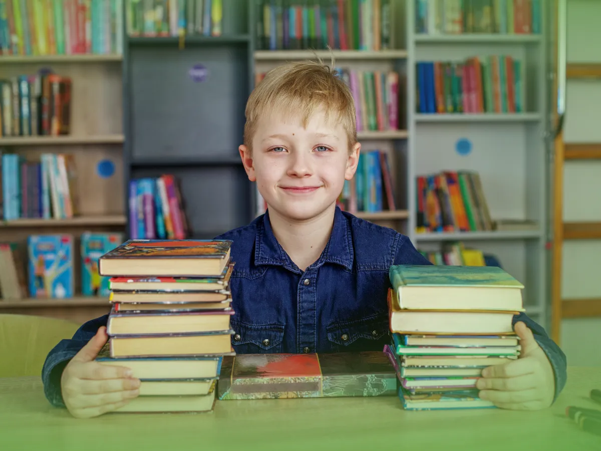 Um menino loiro sorrindo para a câmera, sentado a uma mesa de uma biblioteca com pilhas de Explicações de Língua Portuguesa 4.º Ano. Pacote de Horas em ambos os lados dele.