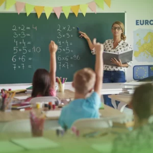 Um professor aponta equações matemáticas num quadro negro numa sala de aula enquanto jovens estudantes levantam as mãos para responder Explicações de Matemática 2.º Ano. Pacote de Horas.