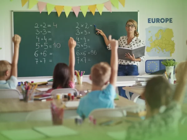 Um professor aponta equações matemáticas num quadro negro numa sala de aula enquanto jovens estudantes levantam as mãos para responder Explicações de Matemática 2.º Ano. Pacote de Horas.