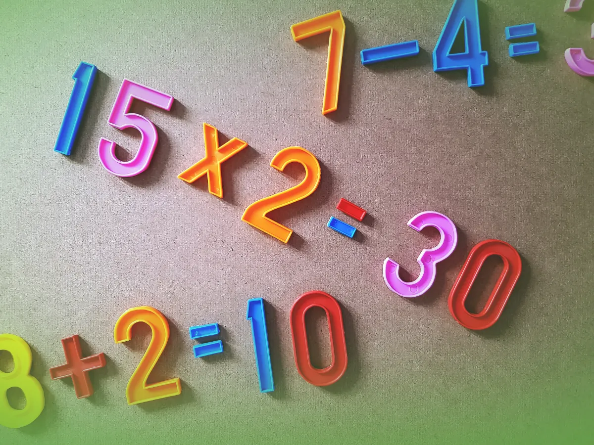 Números magnéticos coloridos sobre um fundo verde exibindo equações matemáticas simples: 15x2=30, 7-4=3, 8+2=10. Explicações de Matemática 3.º Ano . Pacote de Horas