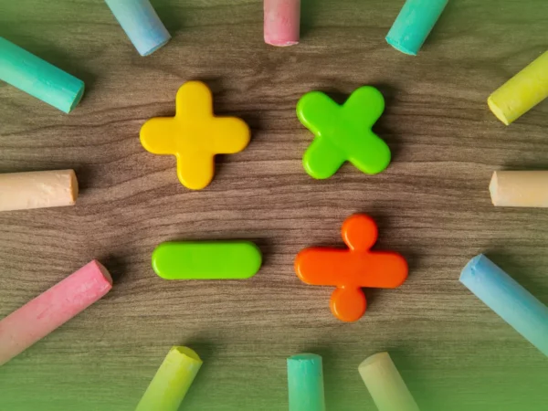 Explicações de Matemática Multicoloridas 4.º Ano . Peças do Pacote de Horas dispostas em círculo sobre uma superfície de madeira, com três peças de giz em formato de sinal de mais em amarelo, verde e laranja no centro.