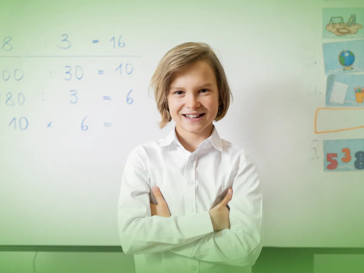 Um menino de camisa branca sorrindo confiante para a câmera com os braços cruzados, em frente a um quadro escolar com problemas de matemática, recebendo Explicações de Matemática 5.º Ano. Pacote de Horas.