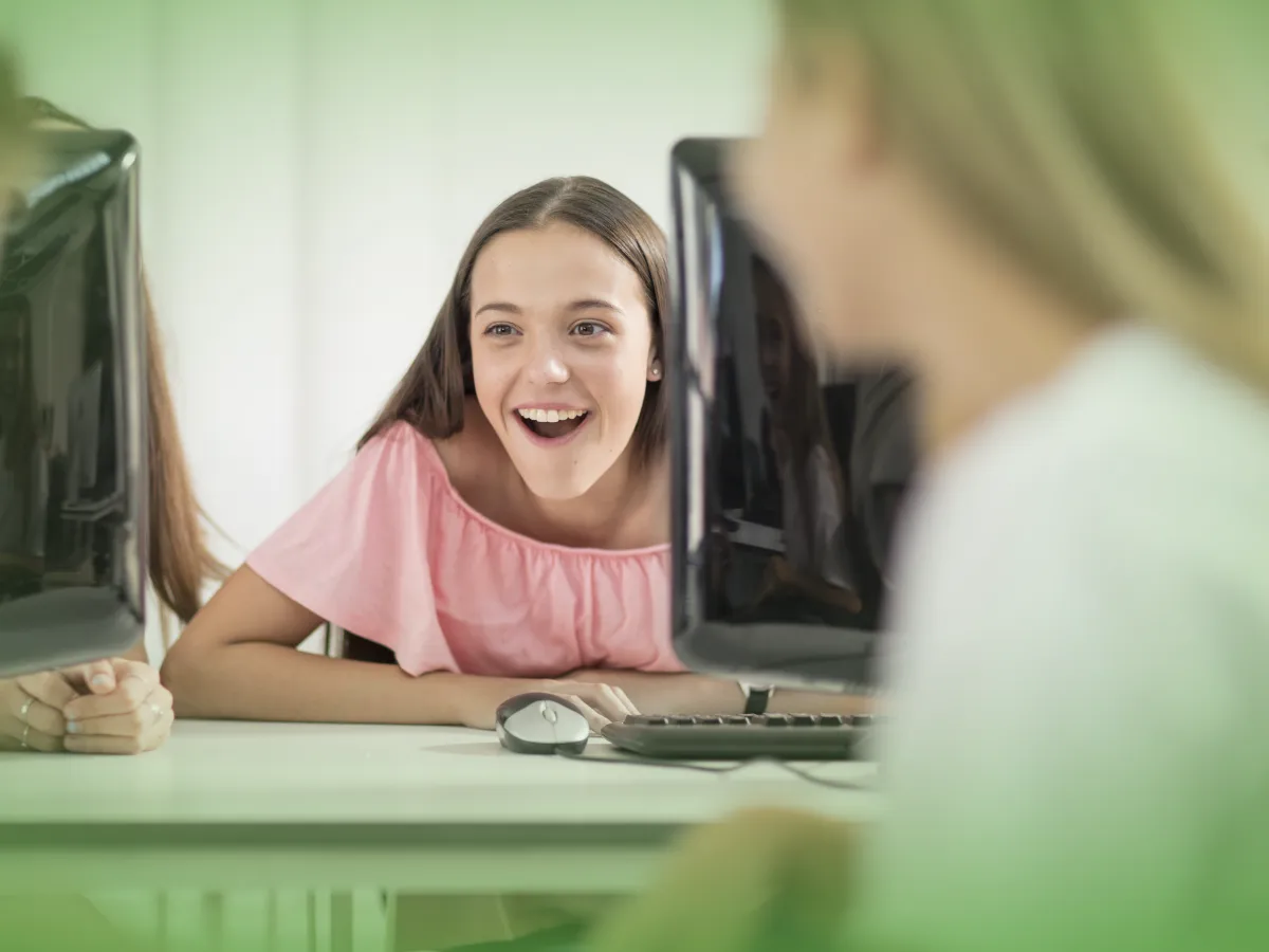 Frase com nome do produto: Duas jovens interagindo alegremente numa mesa de computador em ambiente de sala de aula, uma delas olhando para trás e sorrindo, utilizando as Explicações de Tecnologias de Informação e Comunicação (TIC) 9.º Ano . Pacote de Horas.