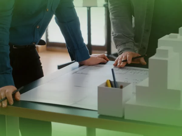 Duas pessoas debruçadas sobre uma mesa examinando com lápis plantas arquitetônicas e Explicações de Urbanismo ONU. Pacote de Horas próximo.