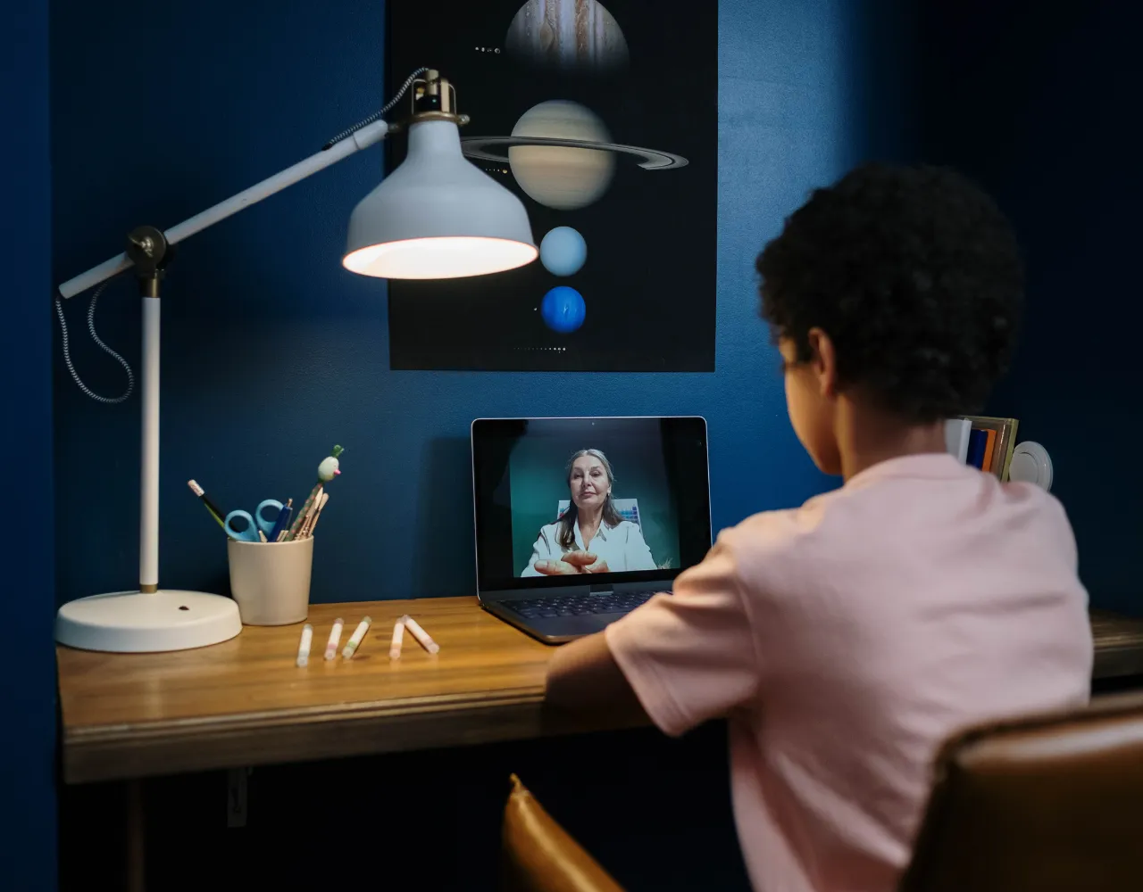Um jovem estudante assiste a uma sessão de sala de aula virtual em um laptop em uma mesa com cartazes educativos e uma lâmpada ao fundo.