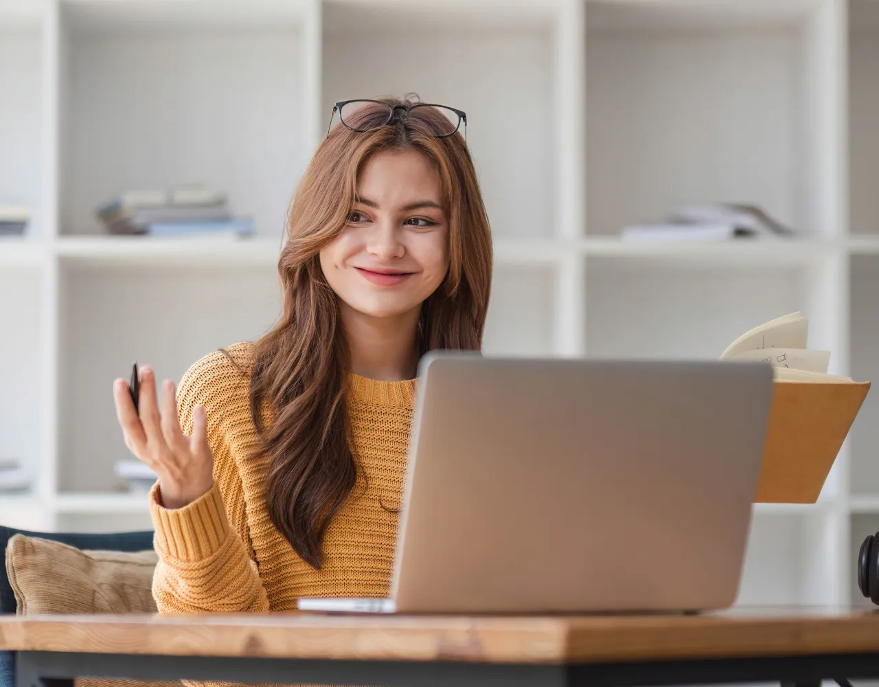 Uma mulher com um suéter amarelo sorri e gesticula enquanto olha para seu laptop em uma sala com prateleiras brancas.