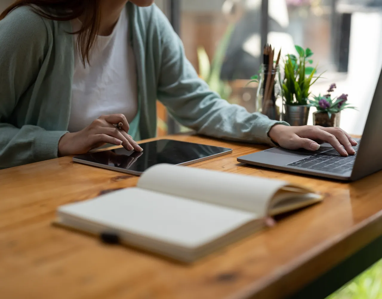 Mulher trabalhando em um laptop e smartphone em uma mesa de madeira com um caderno e uma caneta na frente dela.