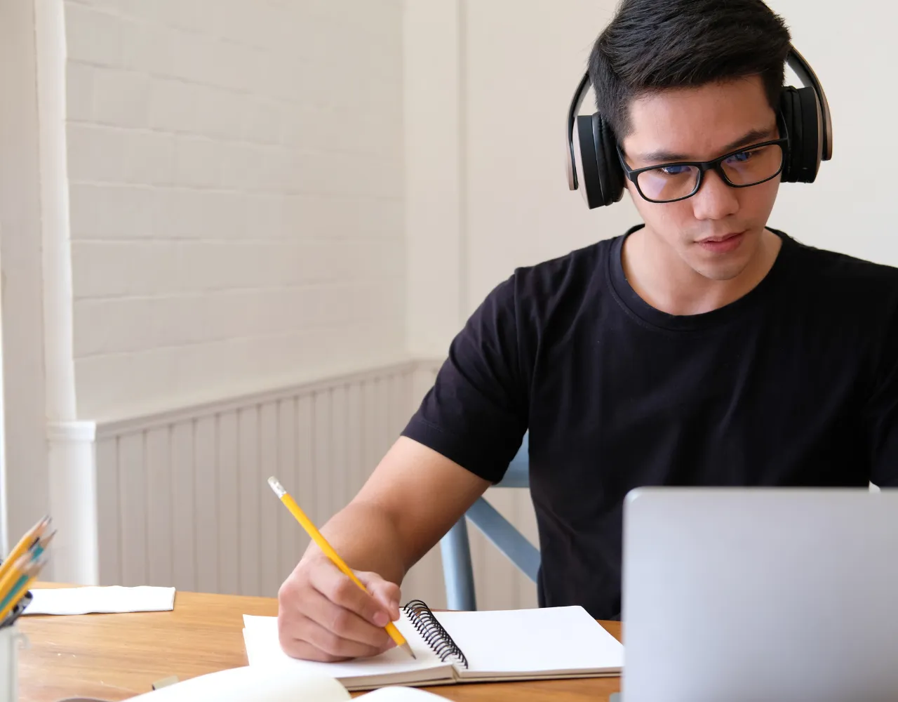 Um jovem focado, usando fones de ouvido e óculos, trabalha em uma mesa com um laptop, fazendo anotações em um caderno.