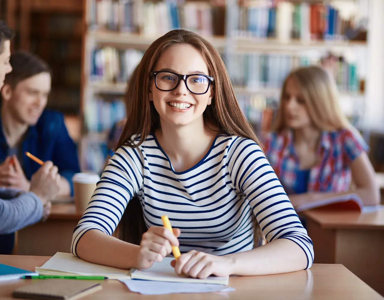 Jovem de óculos e camisa listrada sorri para a câmera enquanto está sentada à mesa em uma biblioteca com colegas ao fundo.
