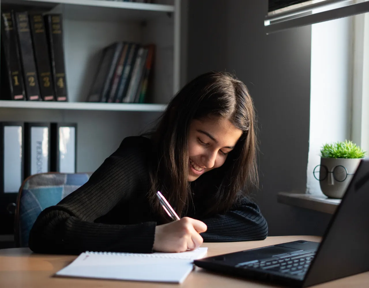 Uma jovem sorrindo enquanto escreve em um caderno em uma mesa, com um laptop aberto ao lado e livros ao fundo.