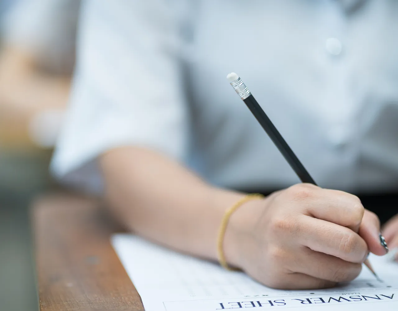 Uma pessoa segura um lápis com a borracha voltada para cima enquanto escreve em uma folha chamada "folha de respostas" sobre uma mesa de madeira.