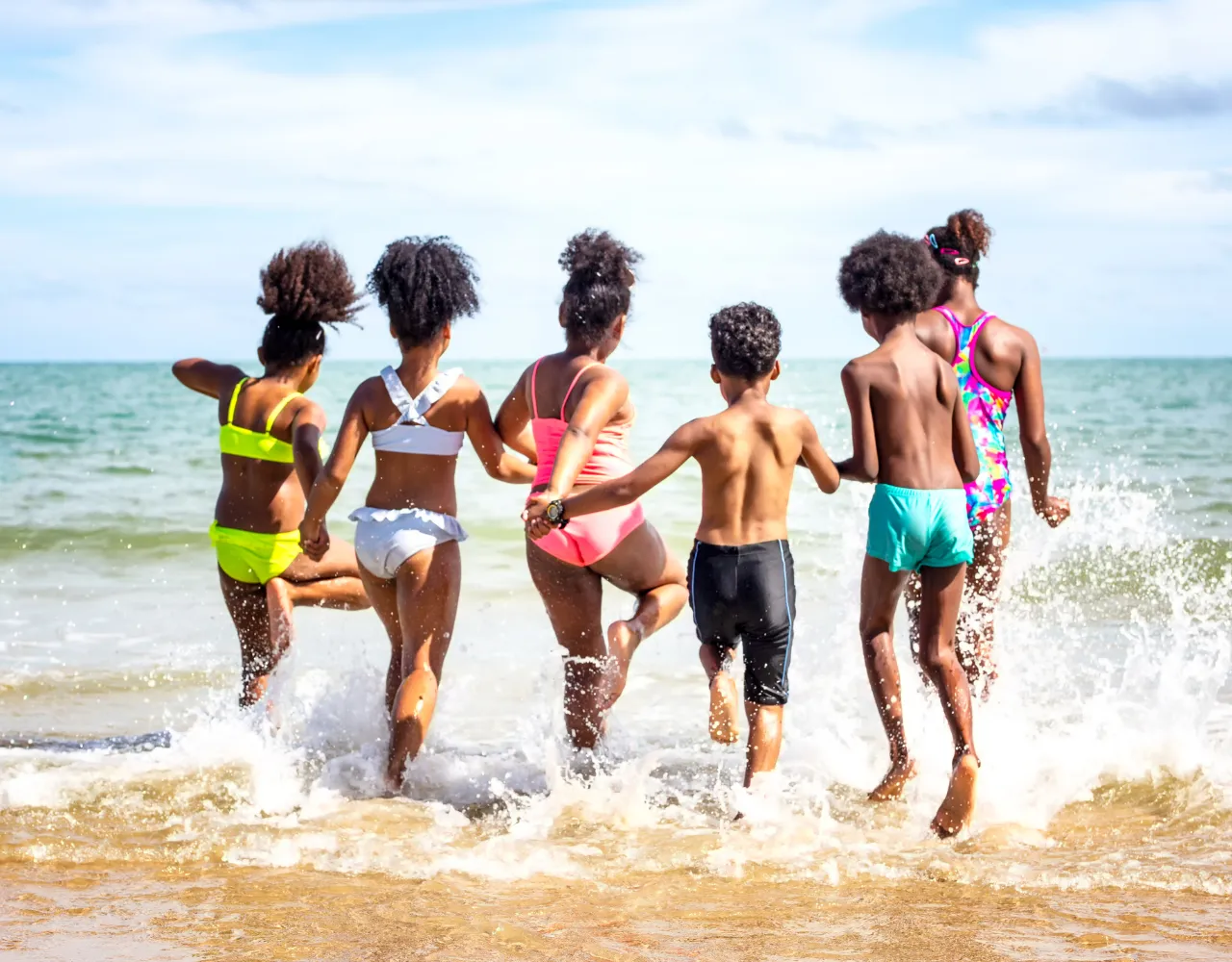 Seis crianças em trajes de banho correm para o mar, de mãos dadas, de costas para a câmera, em um dia ensolarado de praia.