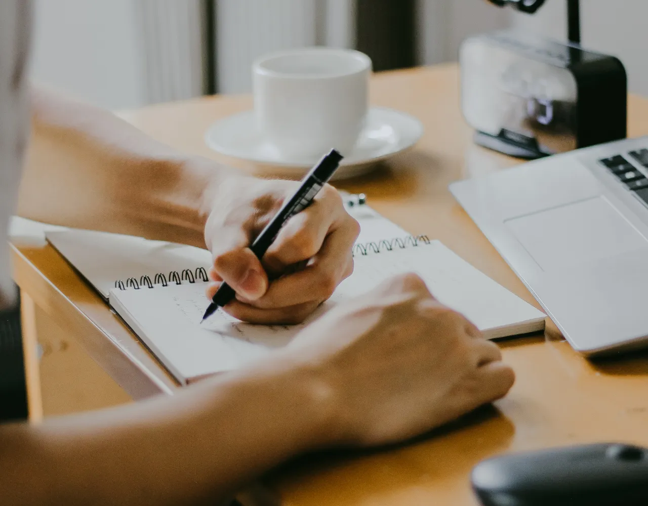 Uma pessoa escreve em um caderno com uma caneta em uma mesa que inclui uma xícara de café, um laptop e um pequeno relógio.