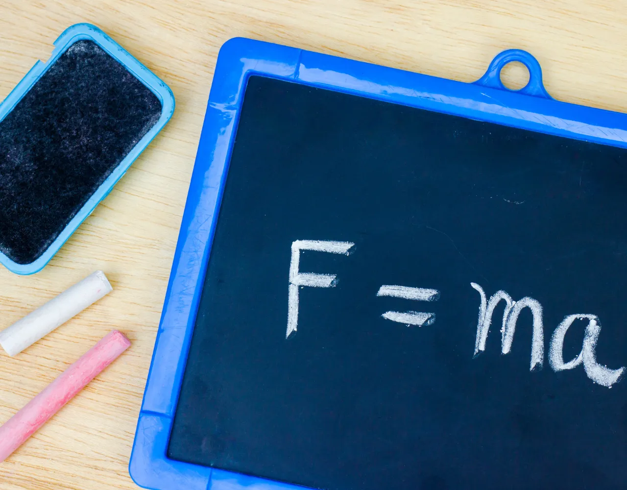 Um quadro negro com moldura azul com a equação "F = ma" escrita em giz branco, um bastão de giz branco e rosa e uma borracha azul colocada sobre uma superfície de madeira.