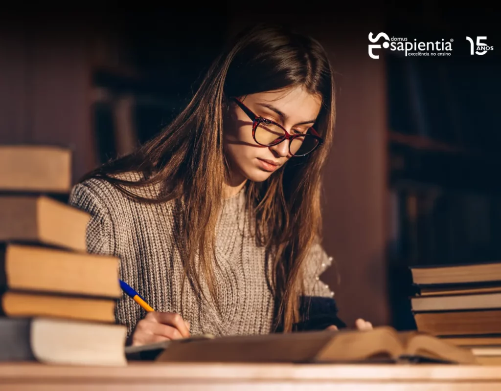 Uma pessoa de óculos escreve em um caderno em uma mesa cercada por livros. O logotipo da "Sapientia" está visível no canto superior direito.