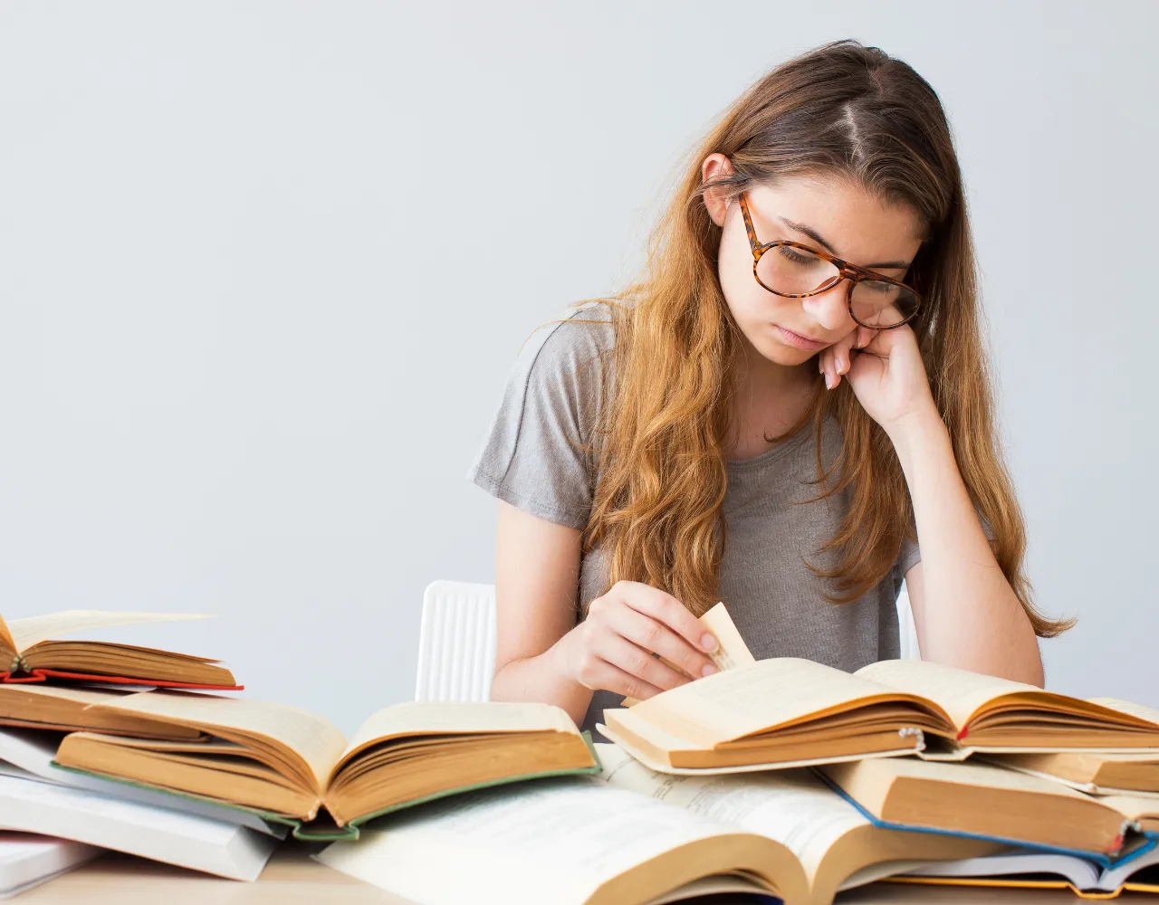 Uma jovem com cabelos longos e óculos está sentada a uma mesa, lendo um livro com vários outros livros abertos ao seu redor. Ela parece profundamente absorta em sua leitura.